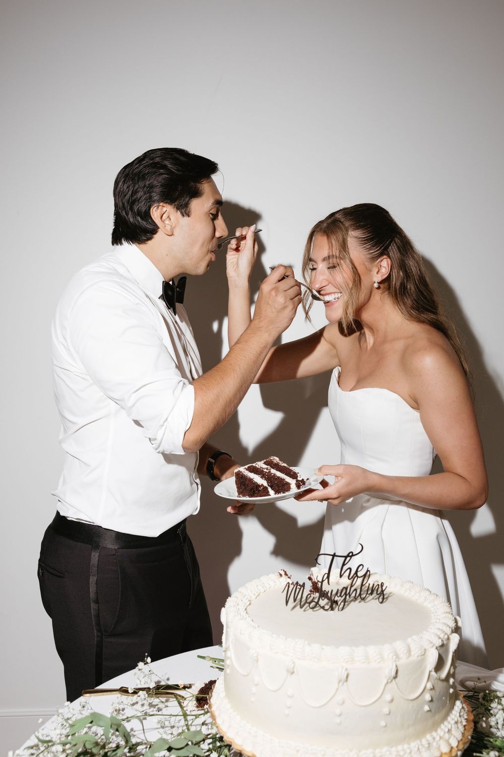 Bride and groom feed each other wedding cake at reception, smiling joyfully beside white tiered cake.