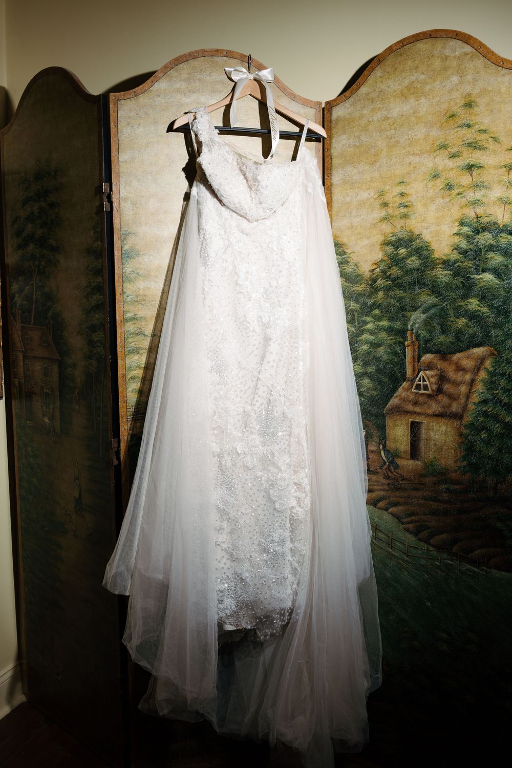Elegant white lace wedding dress hanging against a vintage painted wall with pastoral scenery.