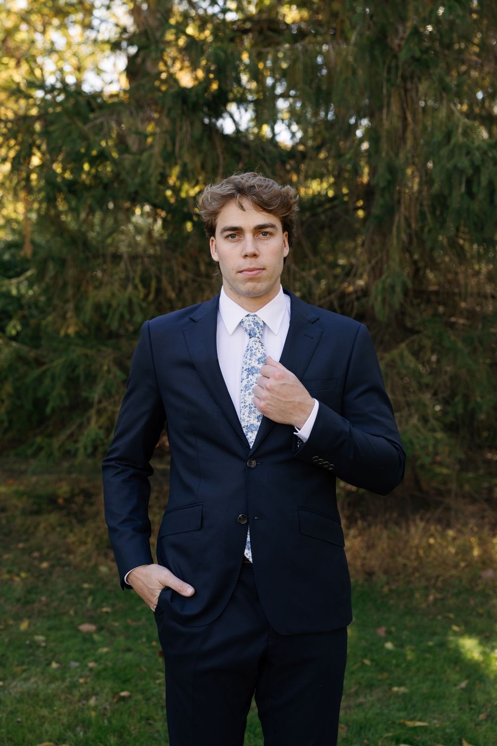 A series of formal portraits showing someone in a navy suit and floral tie posing against a wooded outdoor backdrop.