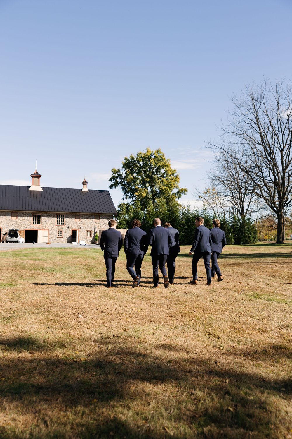 A group of groomsmen in dark suits walk along a grassy hillside path towards a rustic building on a sunny day.