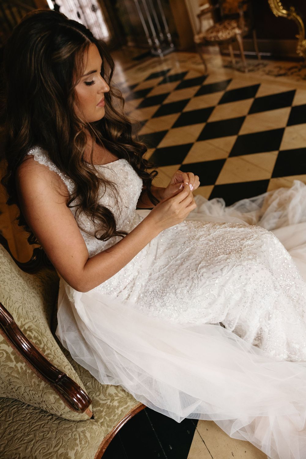 A bride in a white lace wedding dress sits on a black and white checkered floor while holding a bottle.