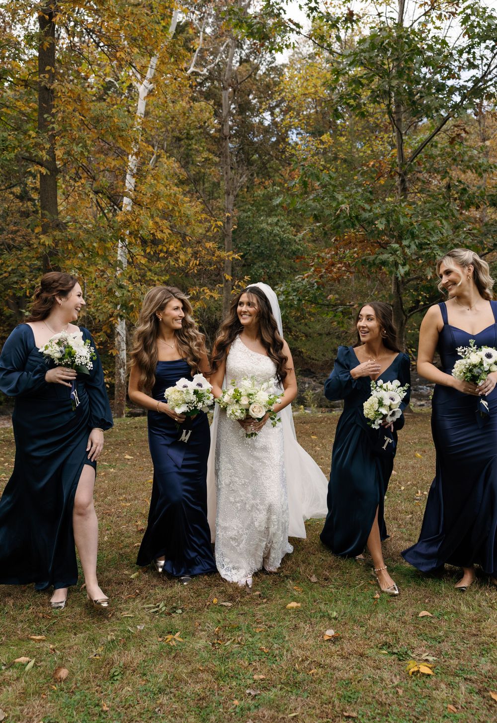 Wedding party laughing and posing together on grass with fall foliage in background.