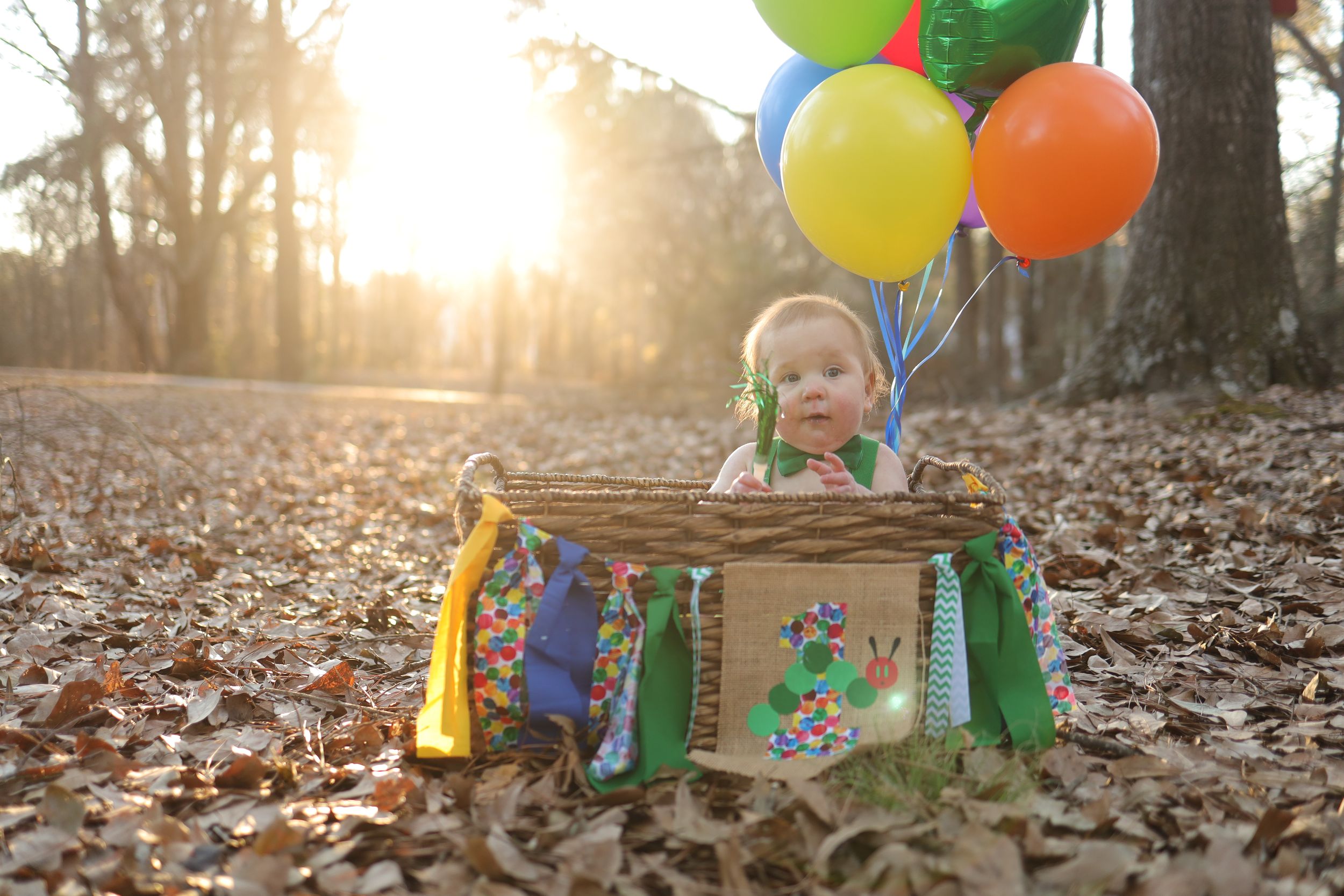 Small child sits in rustic wooden crate with colorful balloons and banners surrounded by autumn leaves at sunset.