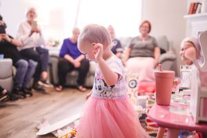 A toddler in a pink tutu dress twirls and dances in a living room during a social gathering.