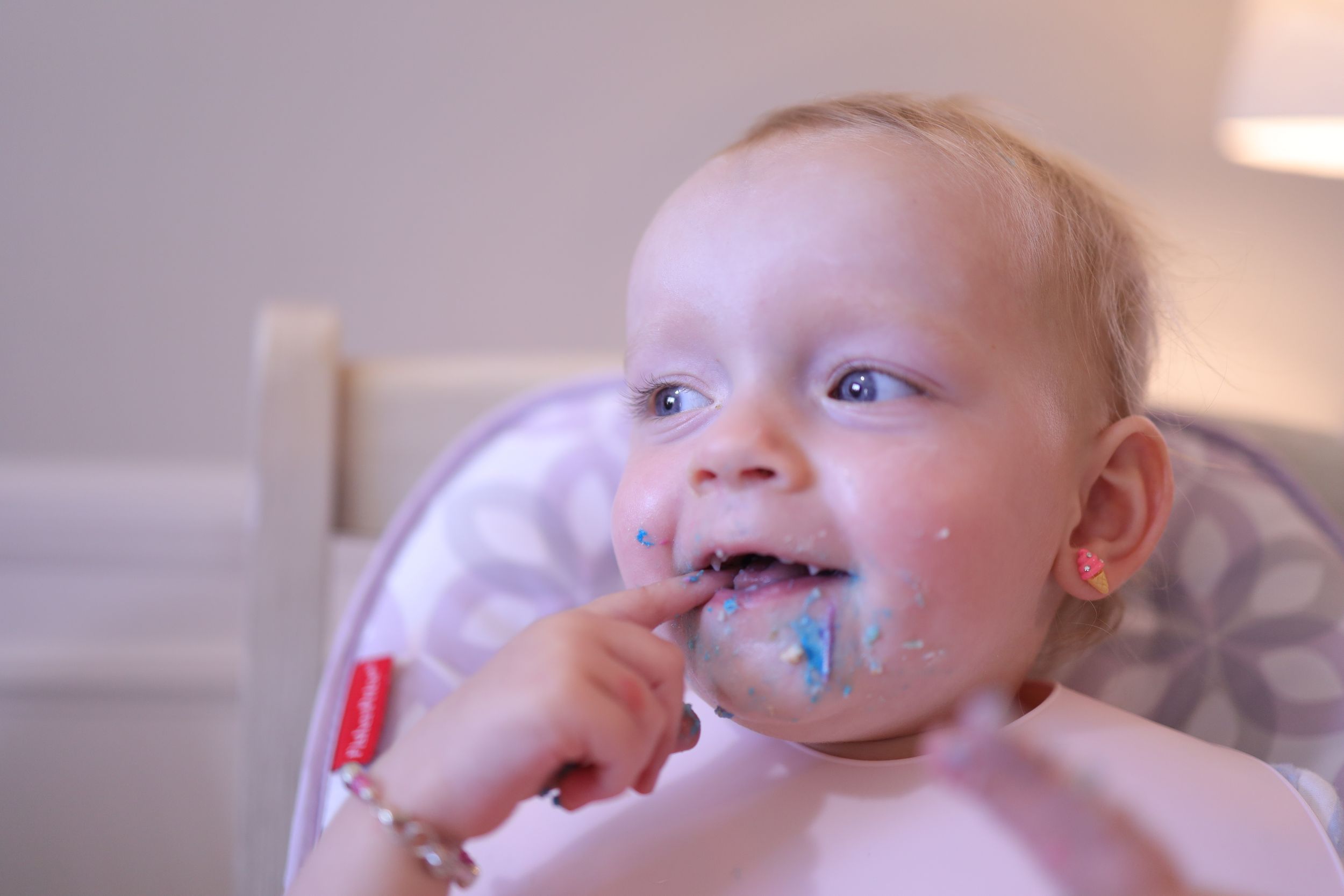 Baby in bib has bright blue food or icing smeared around their mouth while eating in a high chair.