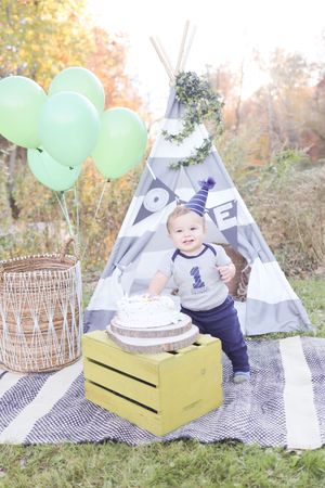 Rustic first birthday setup outdoors with teepee tent, mint balloons, and cake on yellow crate.