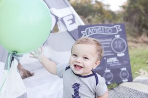 Baby wearing gray outfit with anchor design smiles while reaching for mint green balloon next to chalkboard milestone sign.