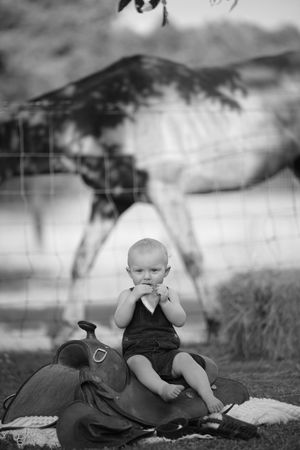 Black and white image showing a child sitting outdoors with rustic saddles in a pastoral setting.