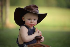 A small child wearing overalls and a brown cowboy hat sits outdoors against a blurred natural background.