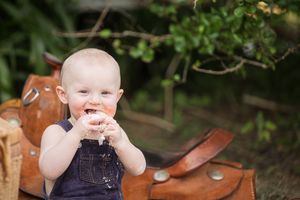 Child in denim overalls sits outdoors smiling and giggling with wooden furniture in the background.