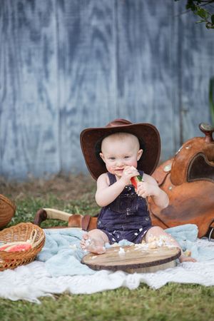 A baby in denim overalls and brown cowboy hat sits on a blanket outdoors with western-themed props and picnic setting.