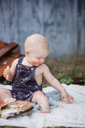 Baby in purple dress plays and makes a mess with cake during cake smash photo shoot on white blanket.