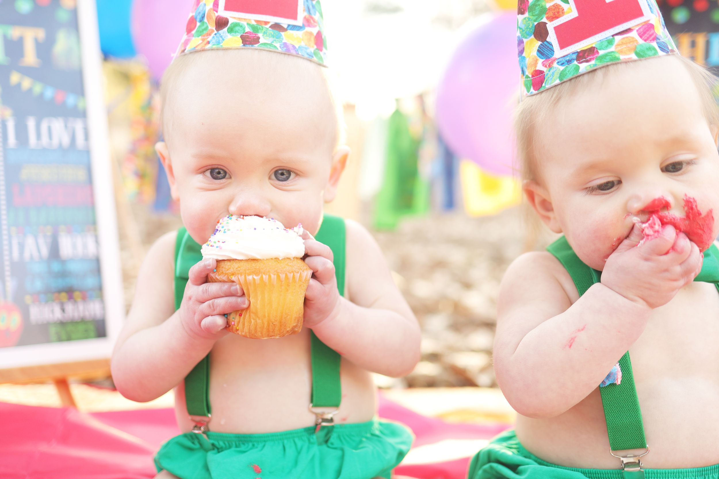 Two babies in green outfits and party hats enjoying frosted cupcakes at a birthday celebration.