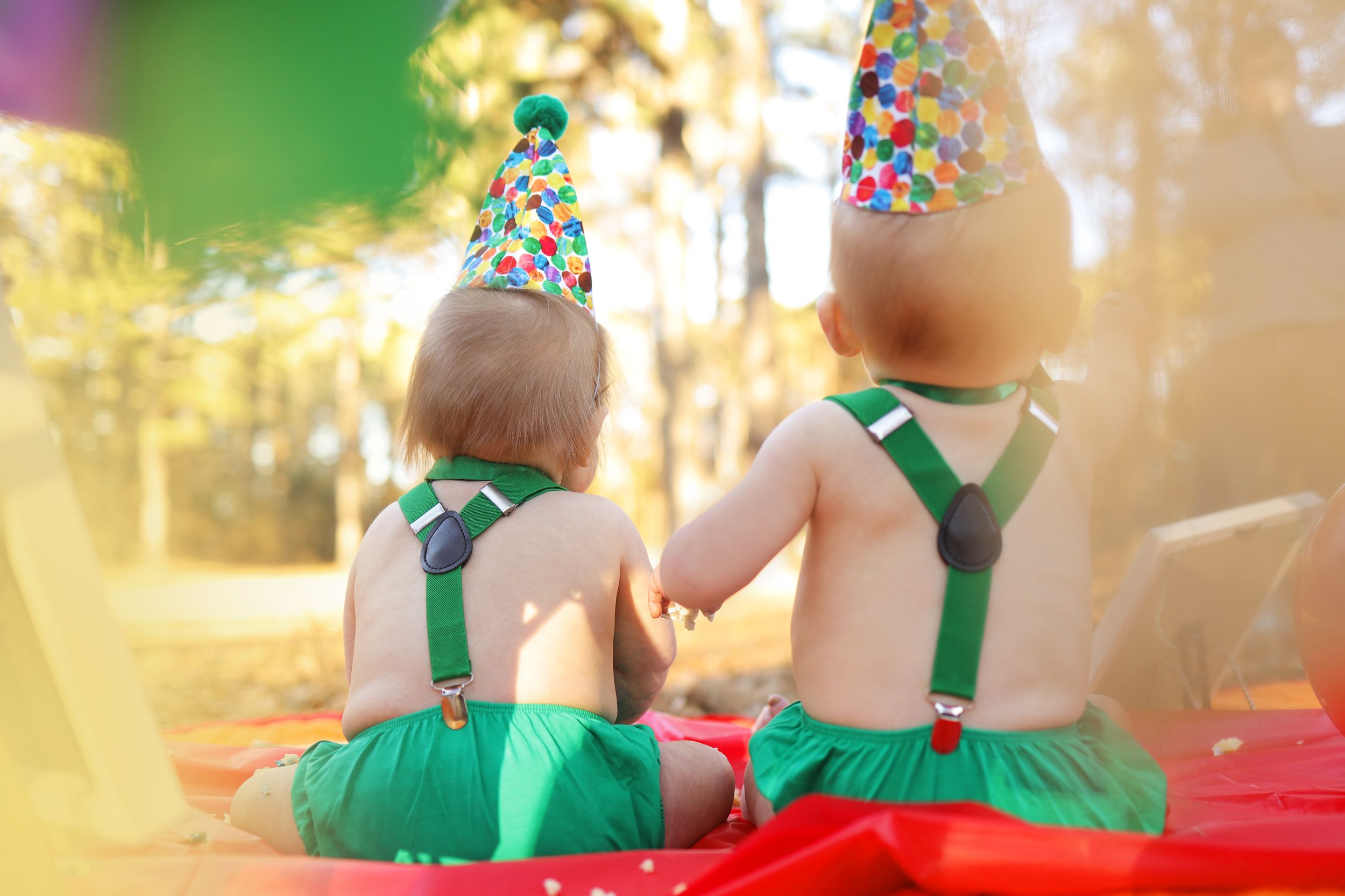 Two children wearing green outfits and party hats sit together at a festive celebration with colorful lighting.