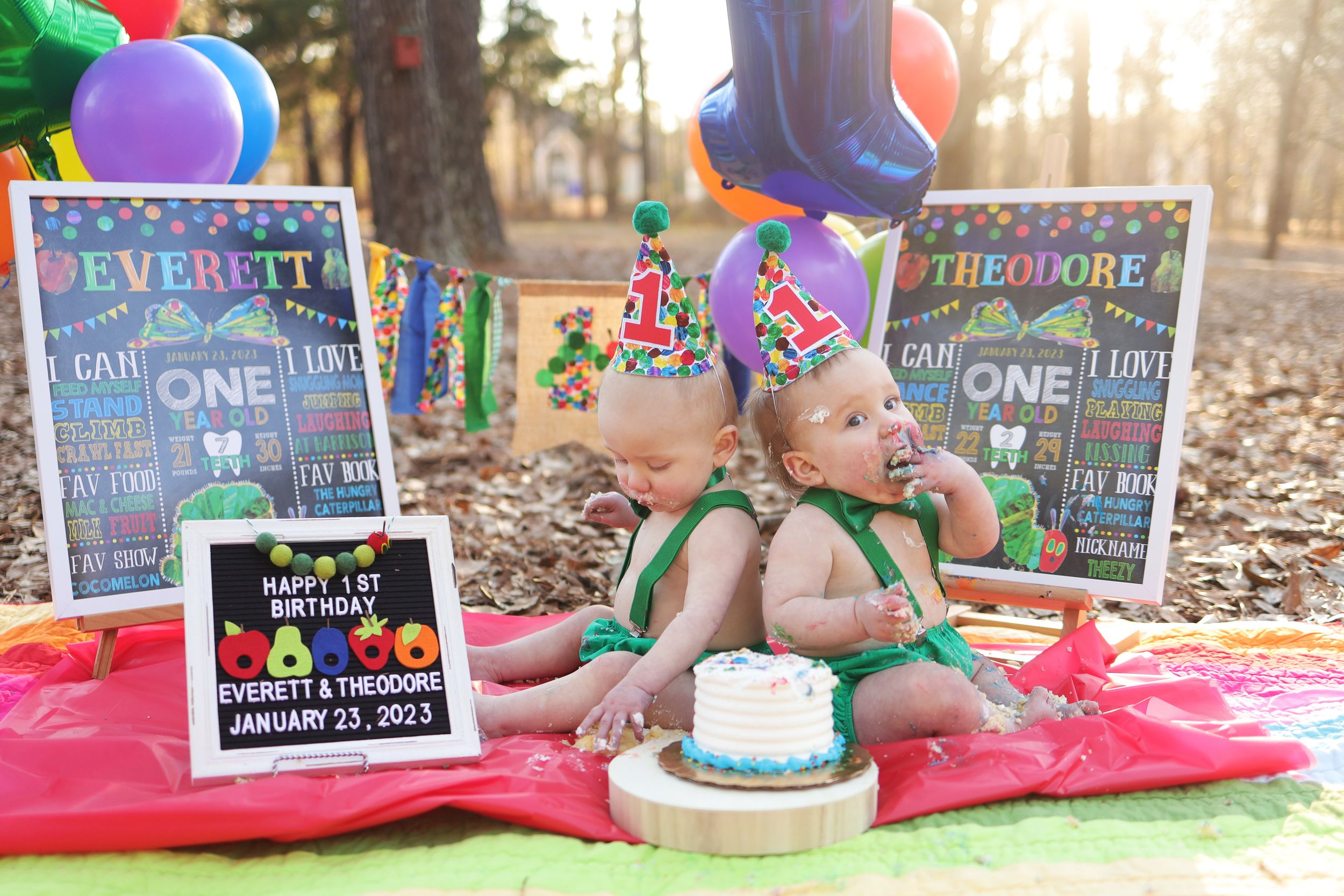 Cake smash first birthday setup outdoors with colorful balloons and chalkboard signs on pink blanket in autumn forest.