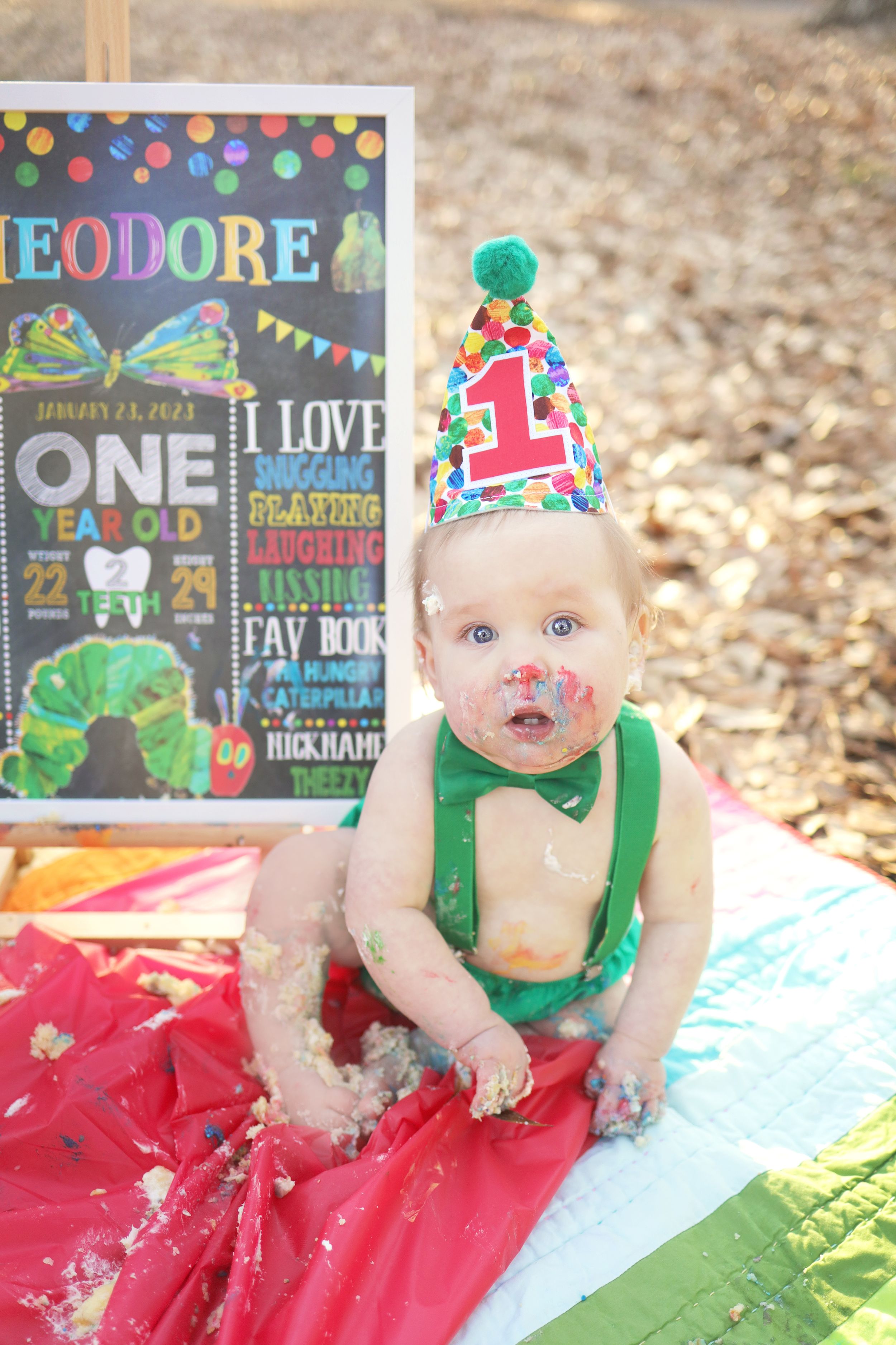 First birthday celebration with chalkboard sign, colorful decorations and baby wearing party hat and green suspenders.
