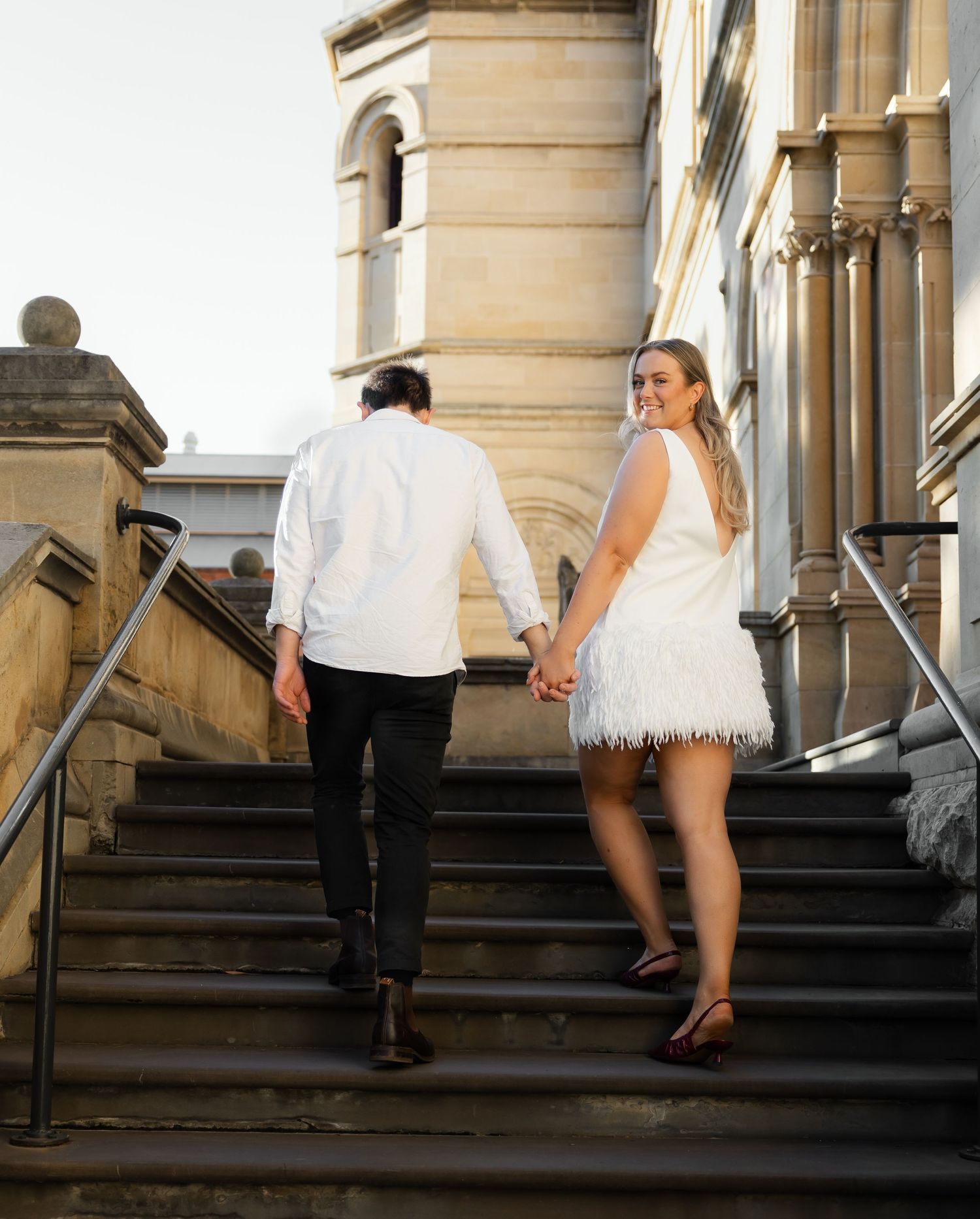 A couple in white attire walking up grand stone steps at a historic building during their wedding celebration.
