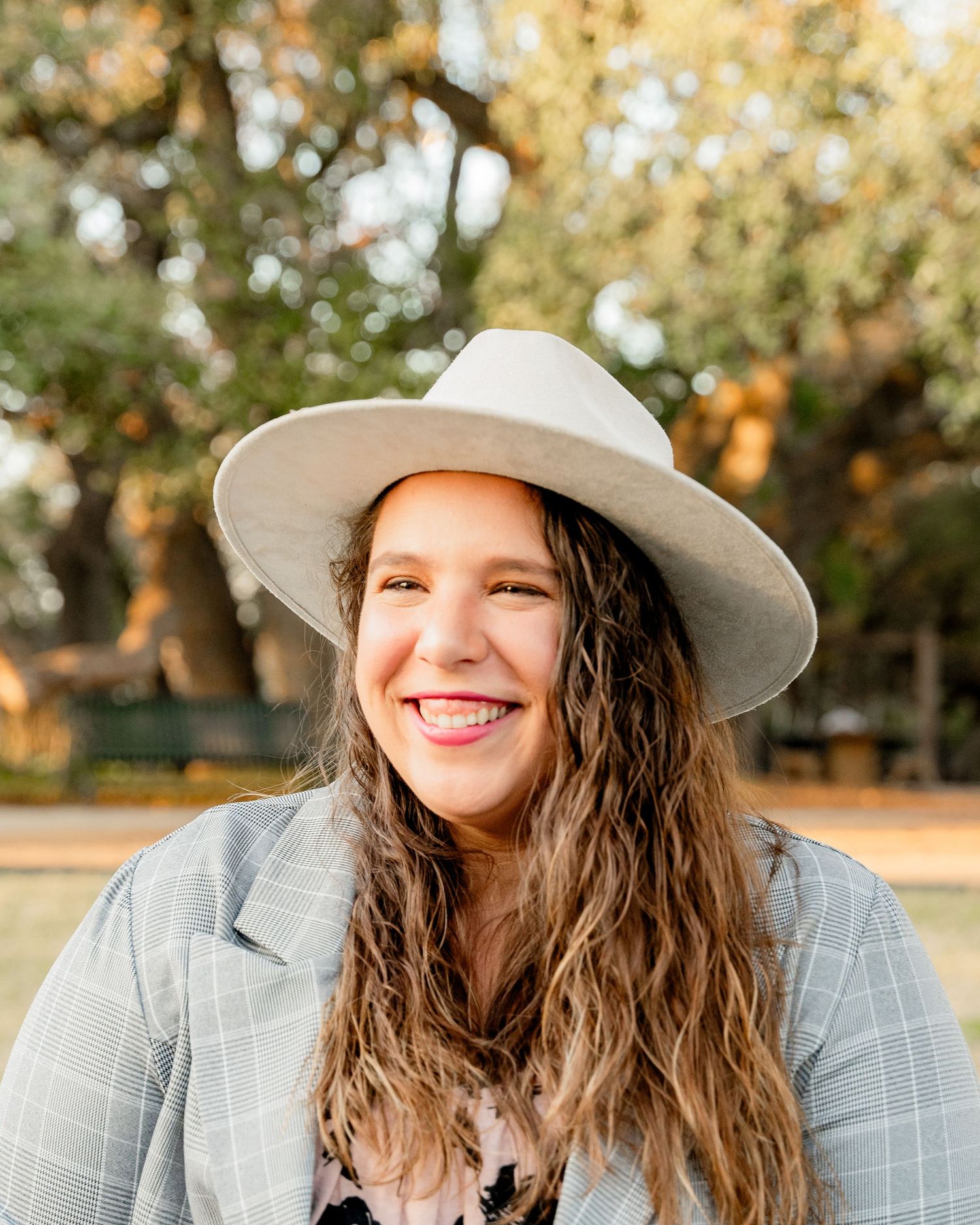 A person in a gray blazer and wide-brimmed hat smiles outdoors during golden hour.