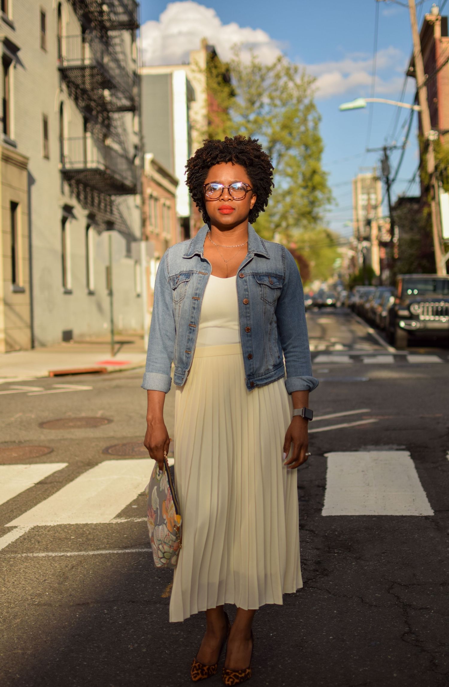 Person in denim jacket and pleated cream dress standing on city street crosswalk in warm afternoon sunlight.