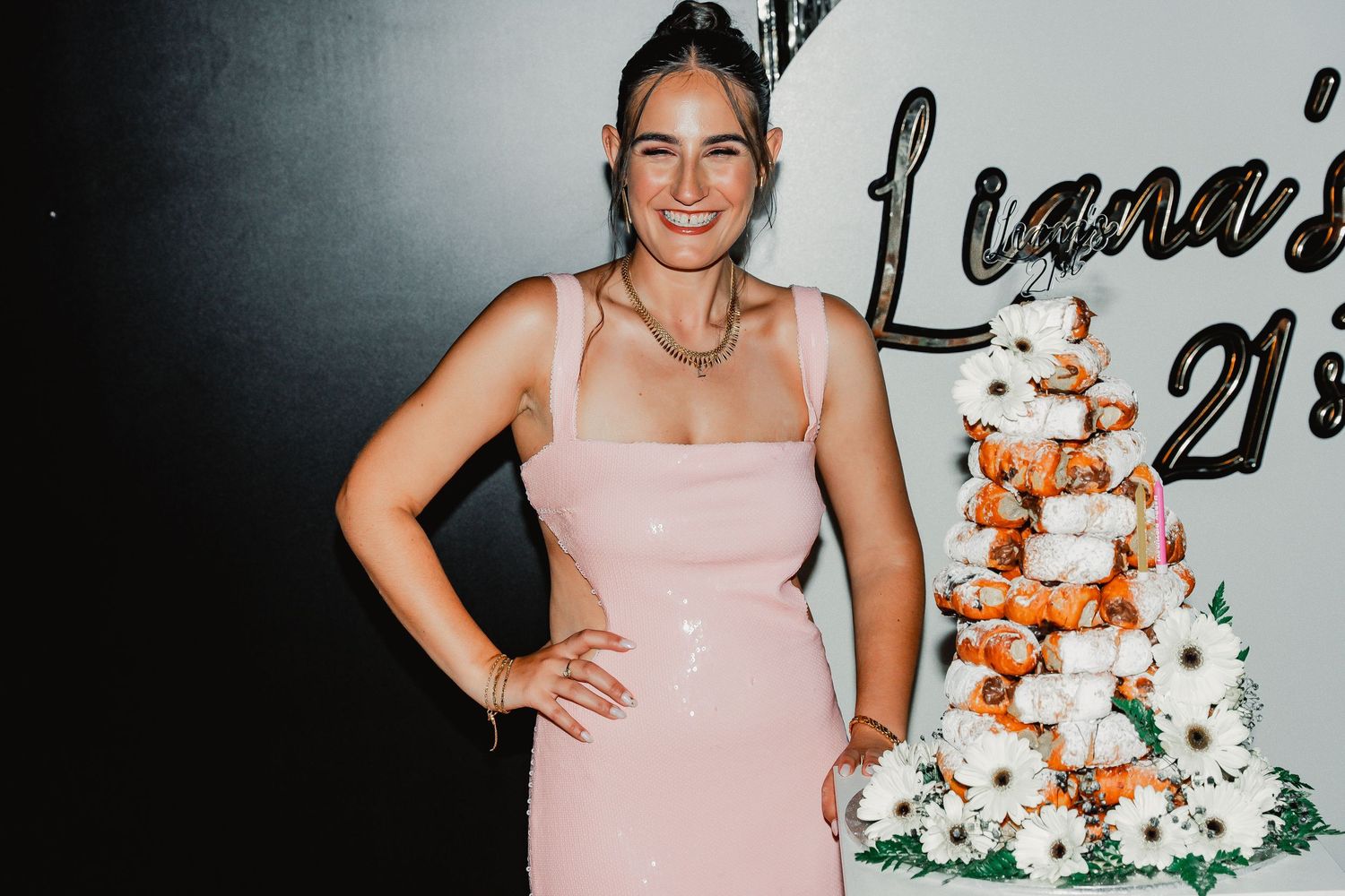 Someone in a pink dress poses with a hand on hip next to an elegant tiered white and copper celebration cake.