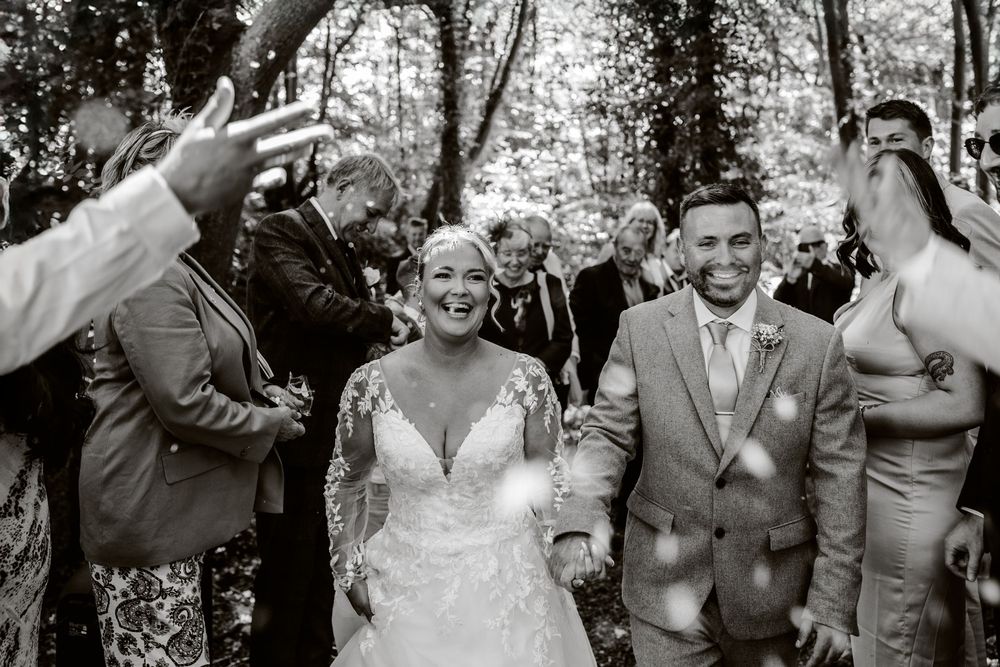 A joyful wedding celebration in a wooded setting as guests toss confetti over a newlywed couple walking down the aisle.
