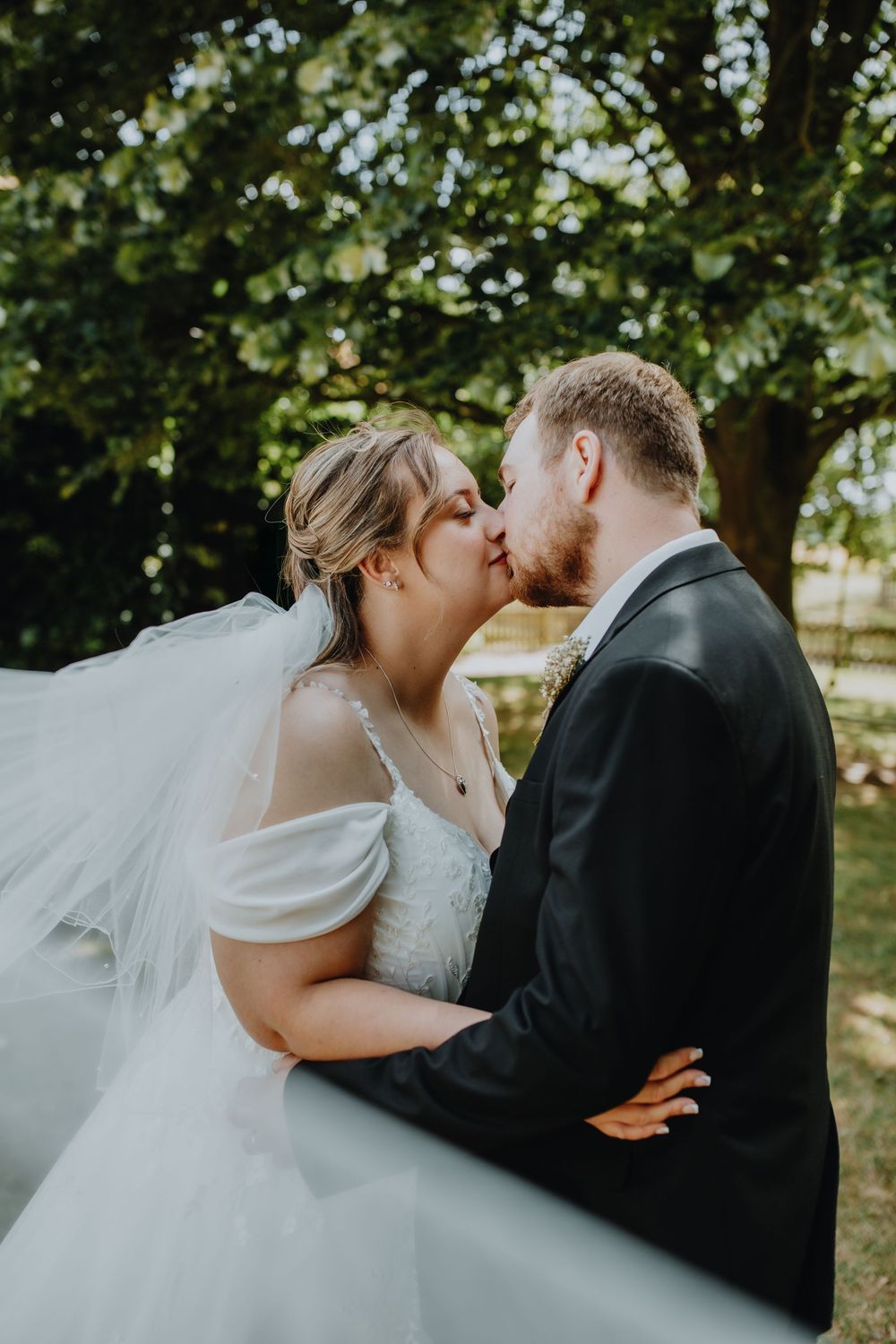 A bride in a flowing white dress and veil shares a romantic kiss with her groom under green trees on their wedding day.
