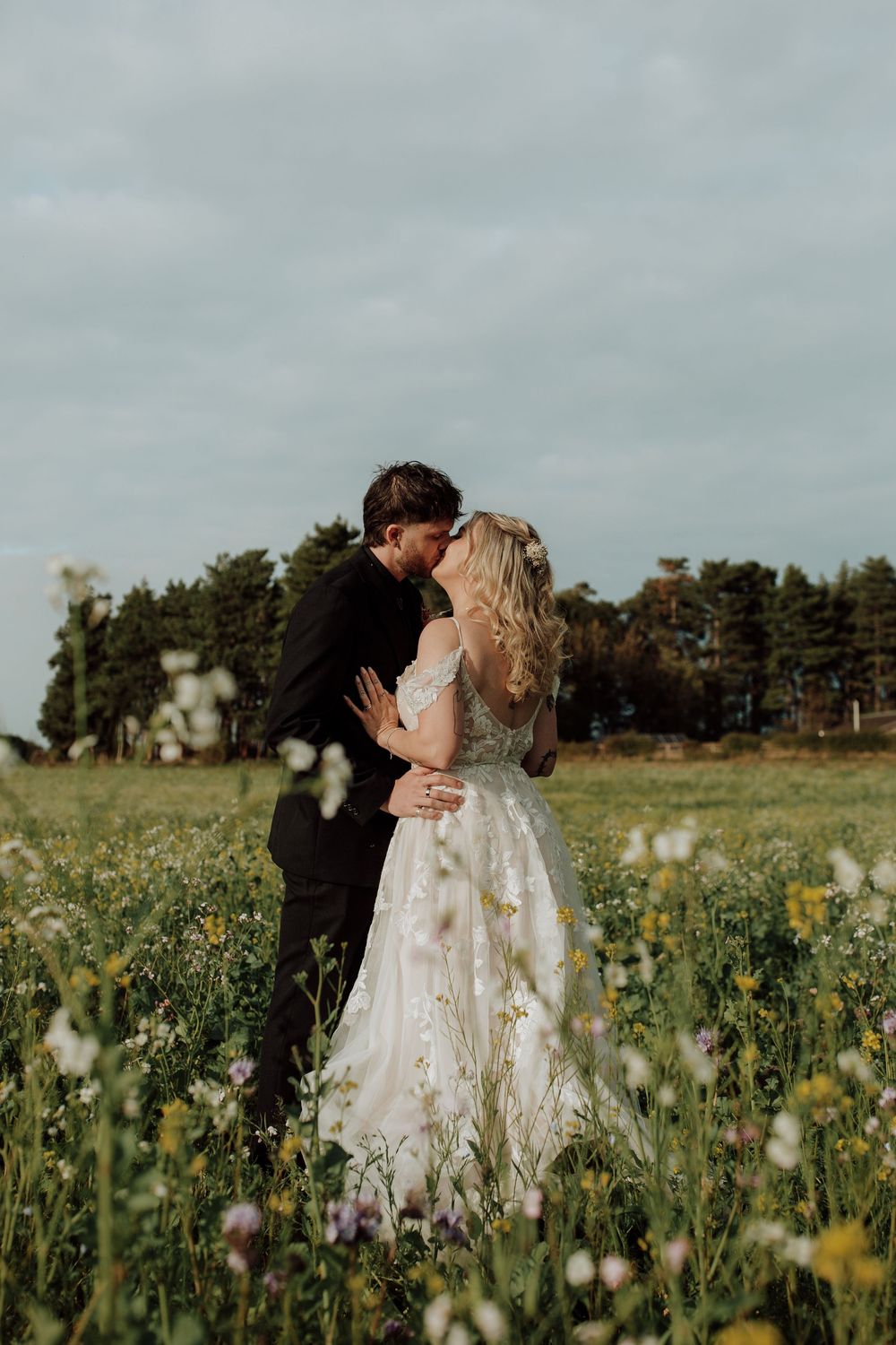 A couple shares a romantic kiss in a wildflower meadow on their wedding day, surrounded by yellow and purple blooms.
