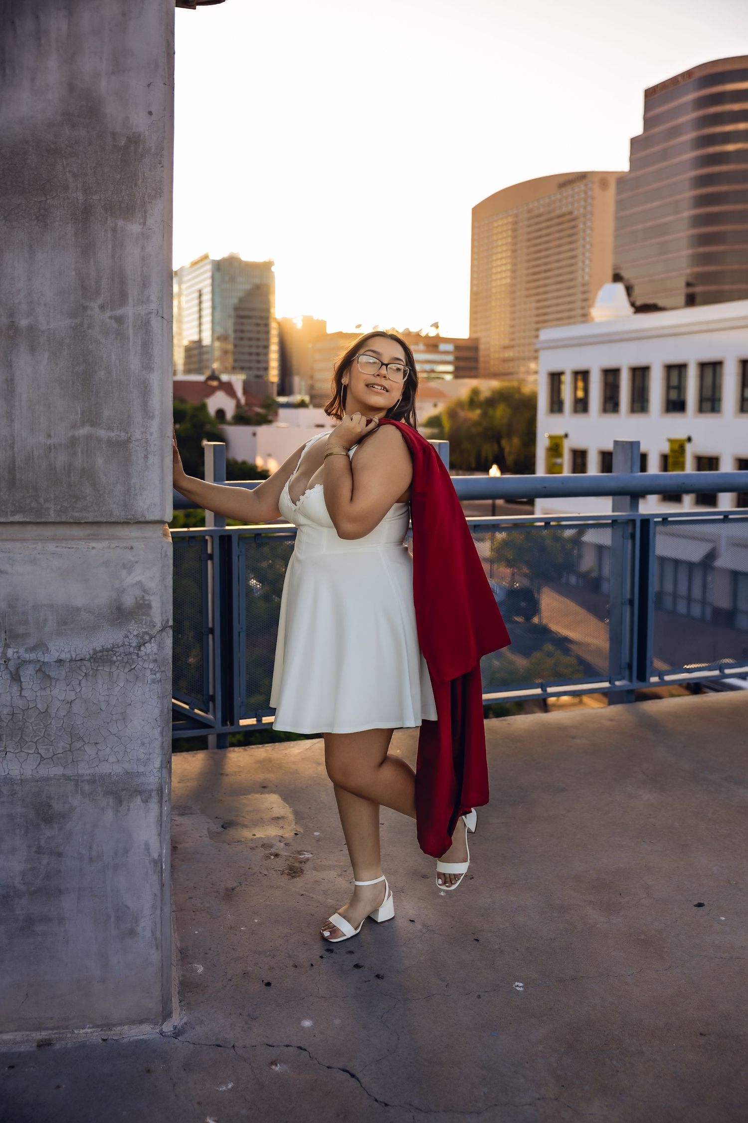 A person in a white sundress and red cardigan poses against a pillar on a rooftop with city buildings in the background.