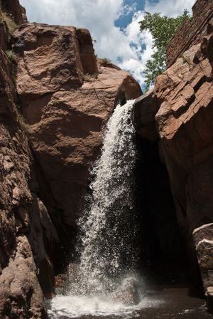 A cascading waterfall streams down between red rock formations under a blue sky with white clouds.