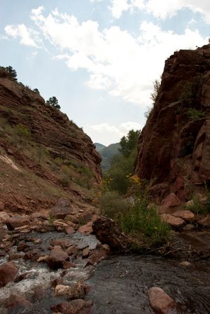 A rushing mountain stream flows between steep red rock canyon walls under a bright blue sky with scattered clouds.