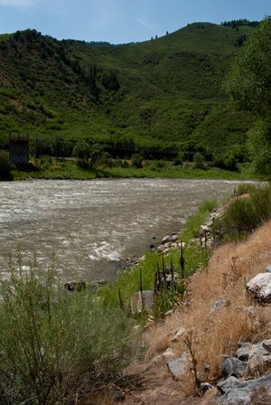 A panoramic view of a rushing river next to an eroded dirt bank with green hills in the background.
