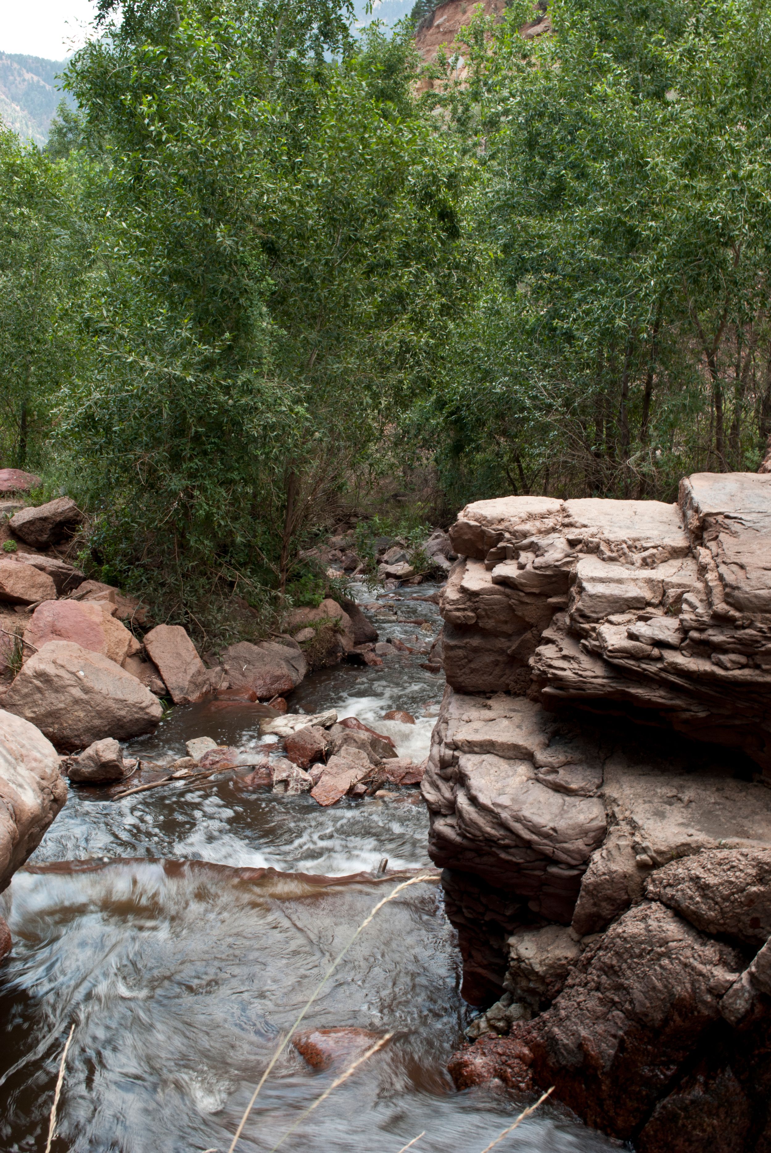 A rushing mountain stream flows through red rock formations surrounded by green foliage in a scenic wilderness setting.
