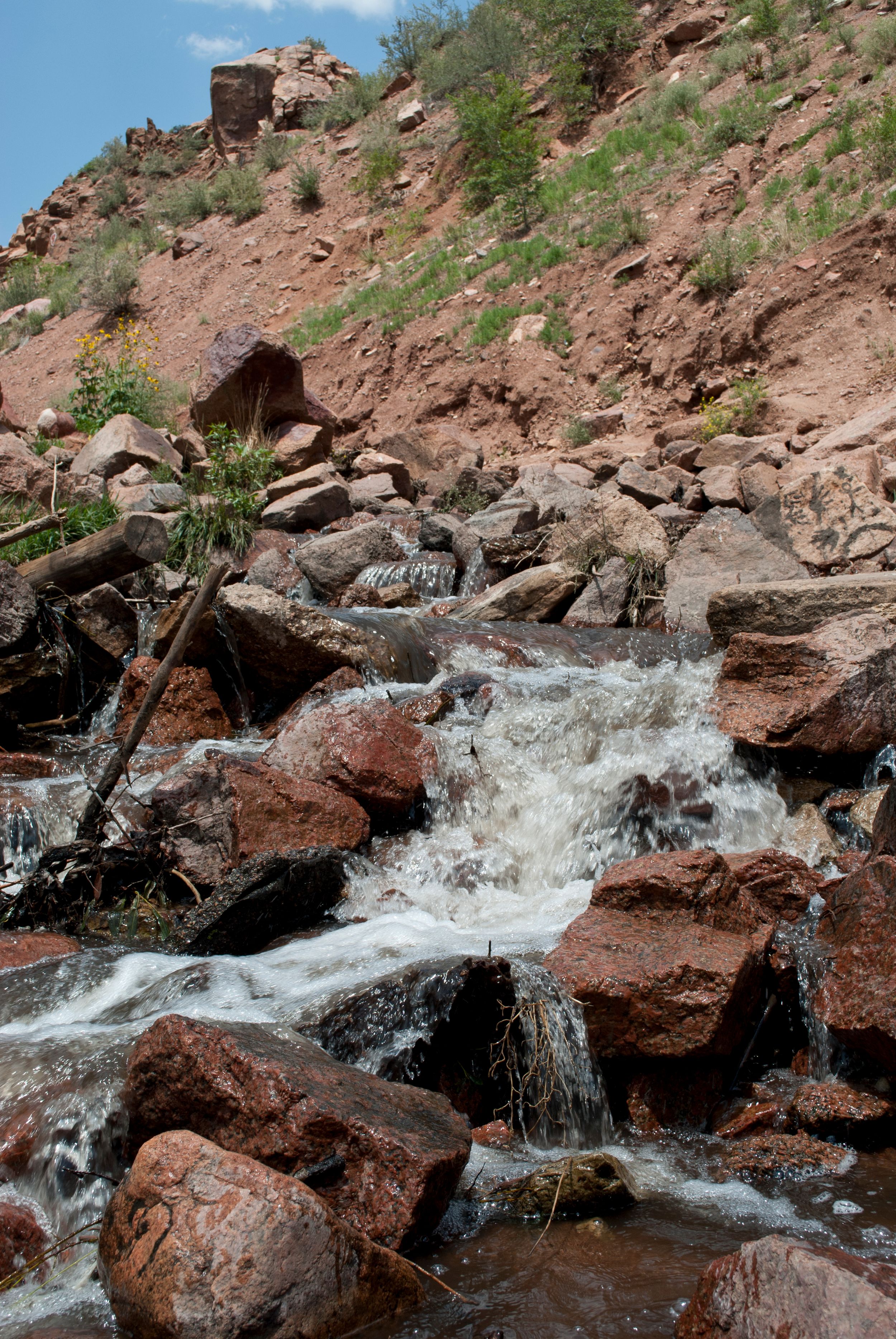 White rushing water cascades down red rocky terrain with scattered vegetation on a mountain slope.