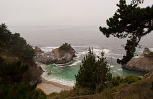 Scenic coastal view of McWay Falls flowing into a turquoise cove surrounded by cliffs and pine trees at Big Sur, California.
