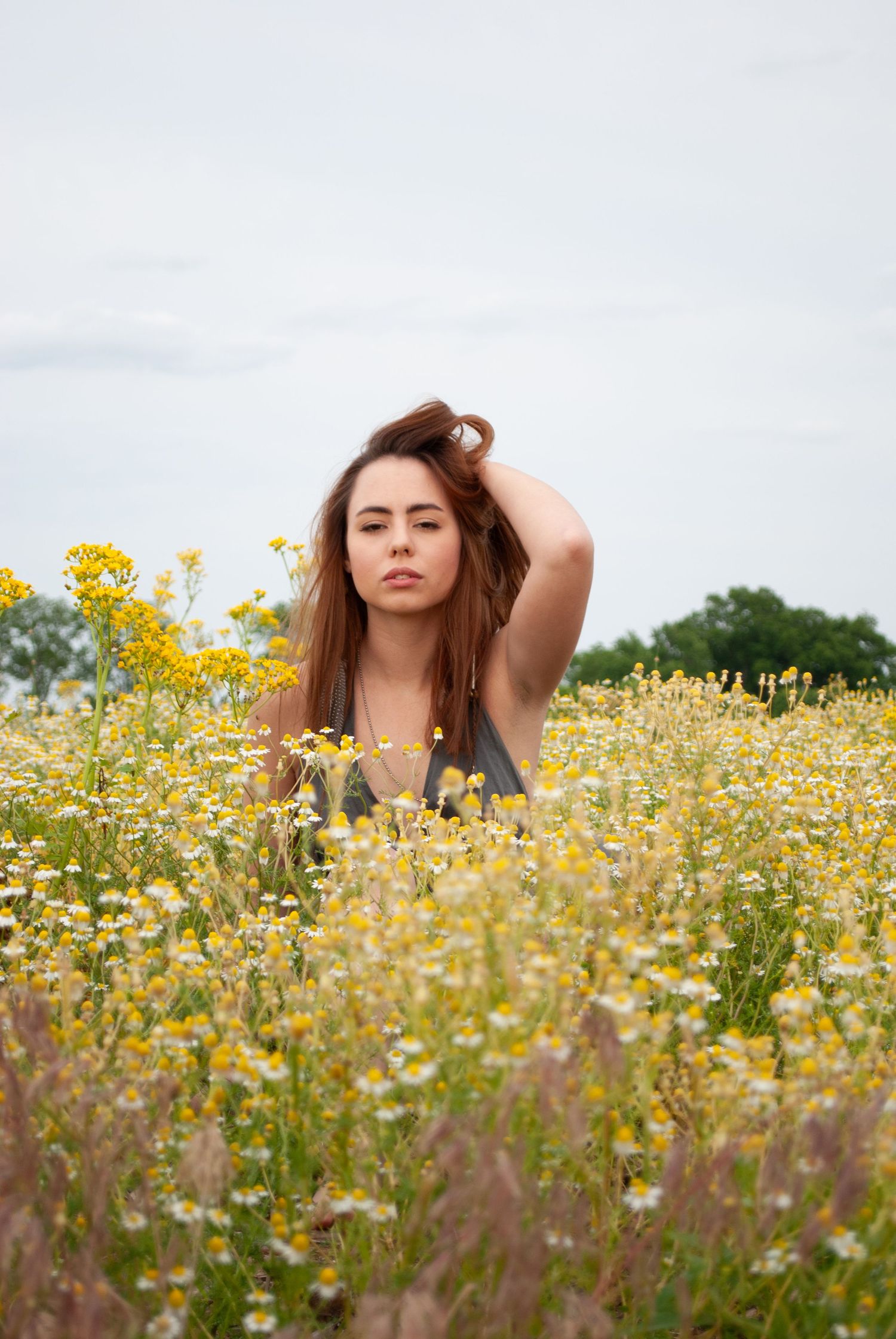A person poses in a field of vibrant yellow wildflowers on an overcast day.