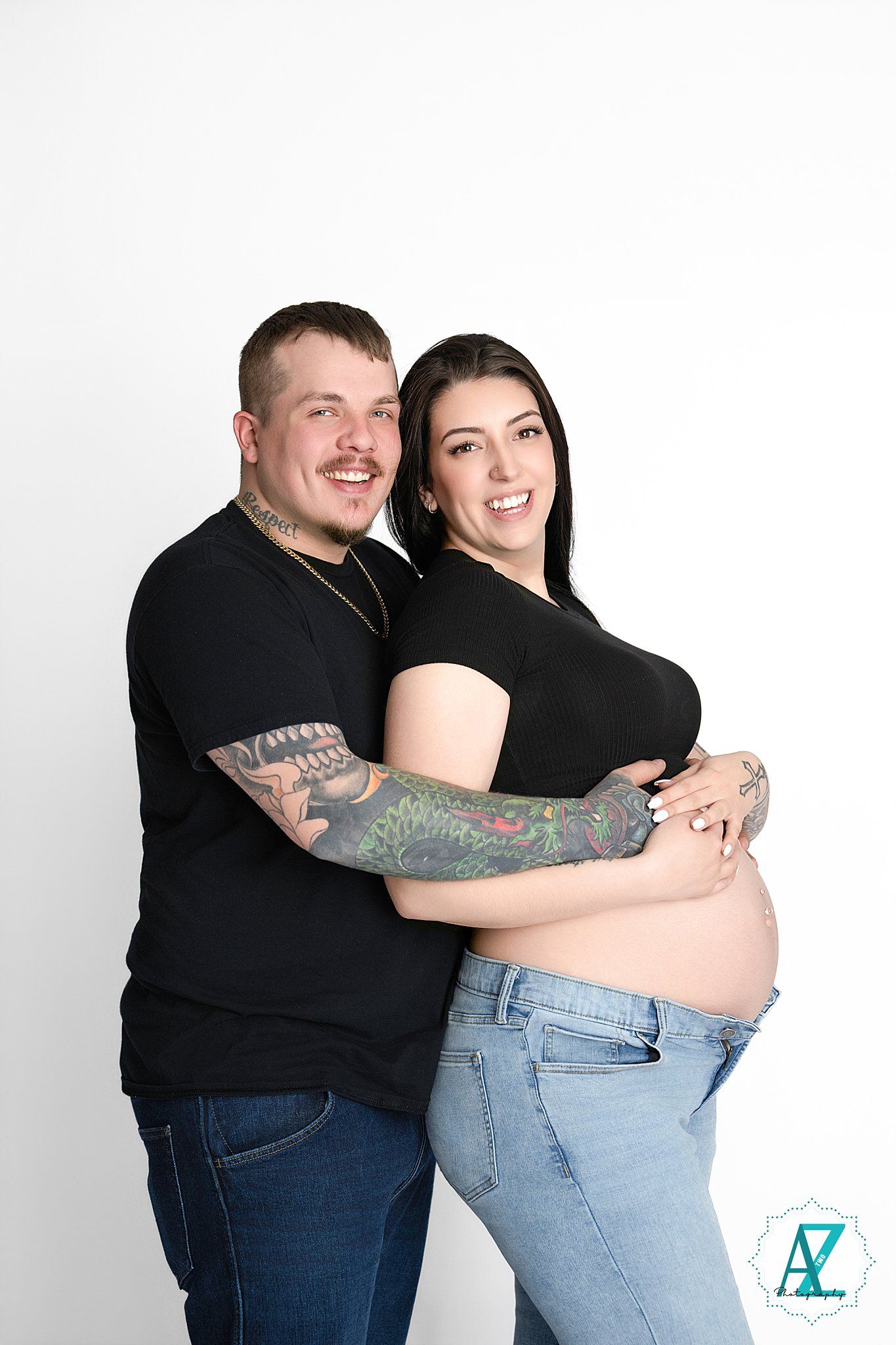 Expectant couple in black tops and jeans embrace in studio maternity photo against white background.