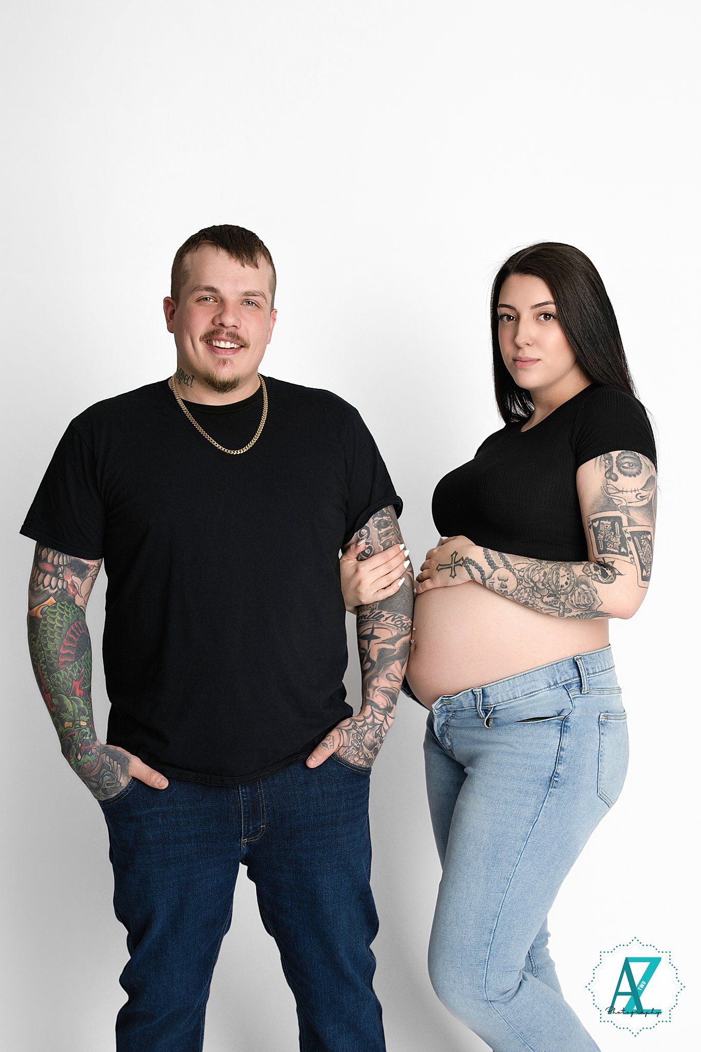 A couple wearing black shirts and jeans pose against a white backdrop, showing off their colorful arm tattoos and baby bump.