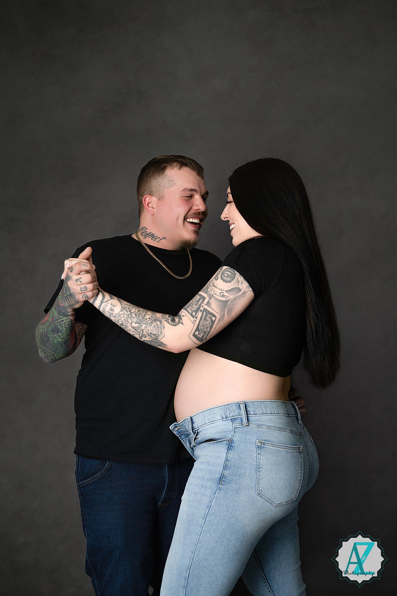 A couple wearing black tops and denim jeans share a happy moment against a dark background, showing off their tattoos.