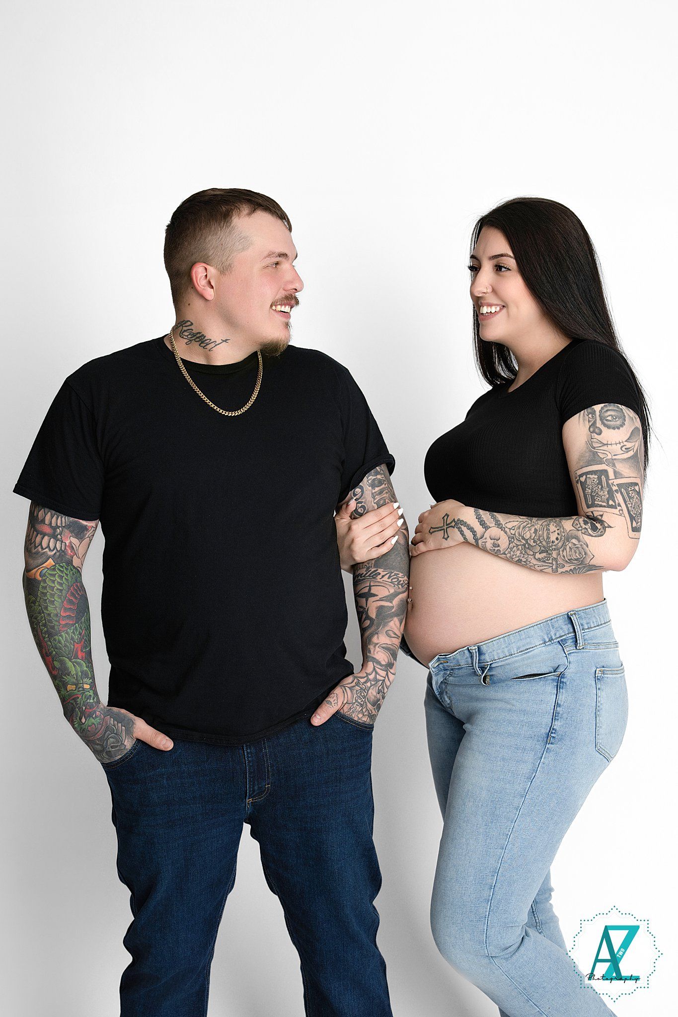 A tattooed couple in casual black tops and jeans pose together against a white background, smiling at each other.