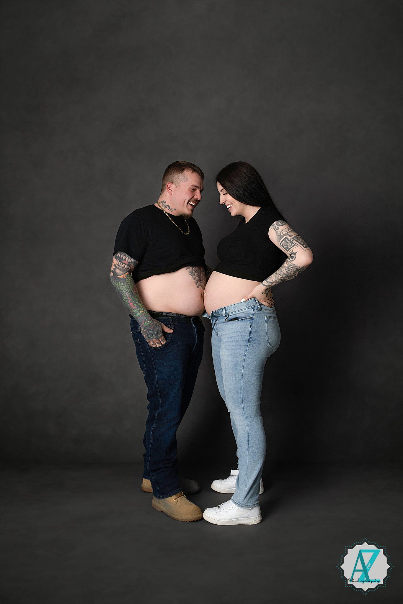 A couple in casual wear facing each other and sharing an intimate moment against a dark backdrop in a studio setting.