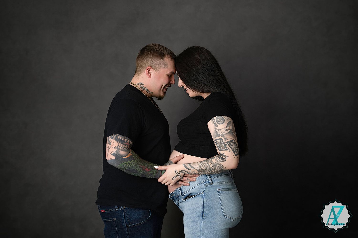 Romantic couple in black shirts with tattoos share an intimate forehead-touching moment against dark backdrop.