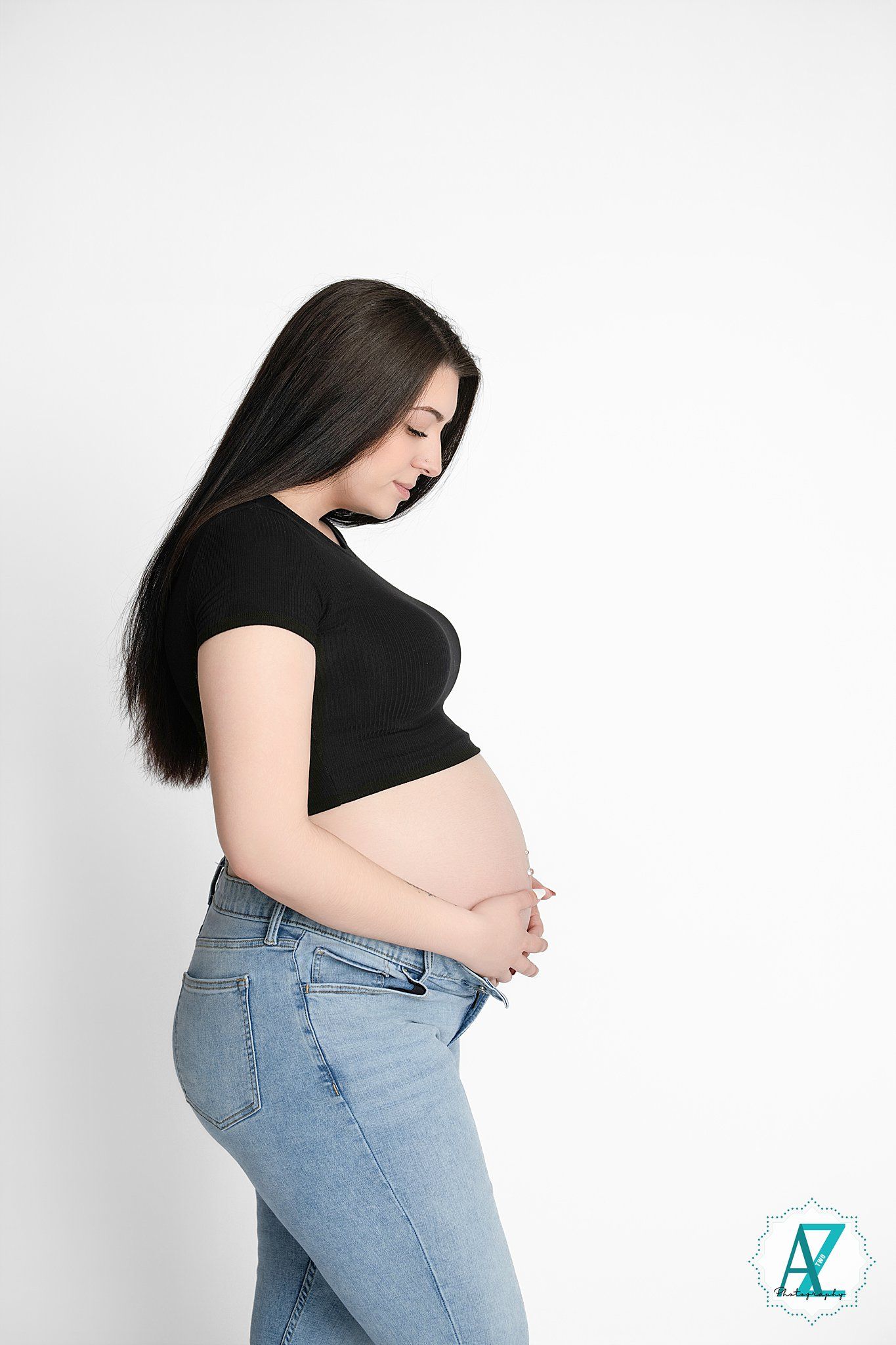 Side view of an expecting mother in a black top and jeans posing against a white background during pregnancy photoshoot.