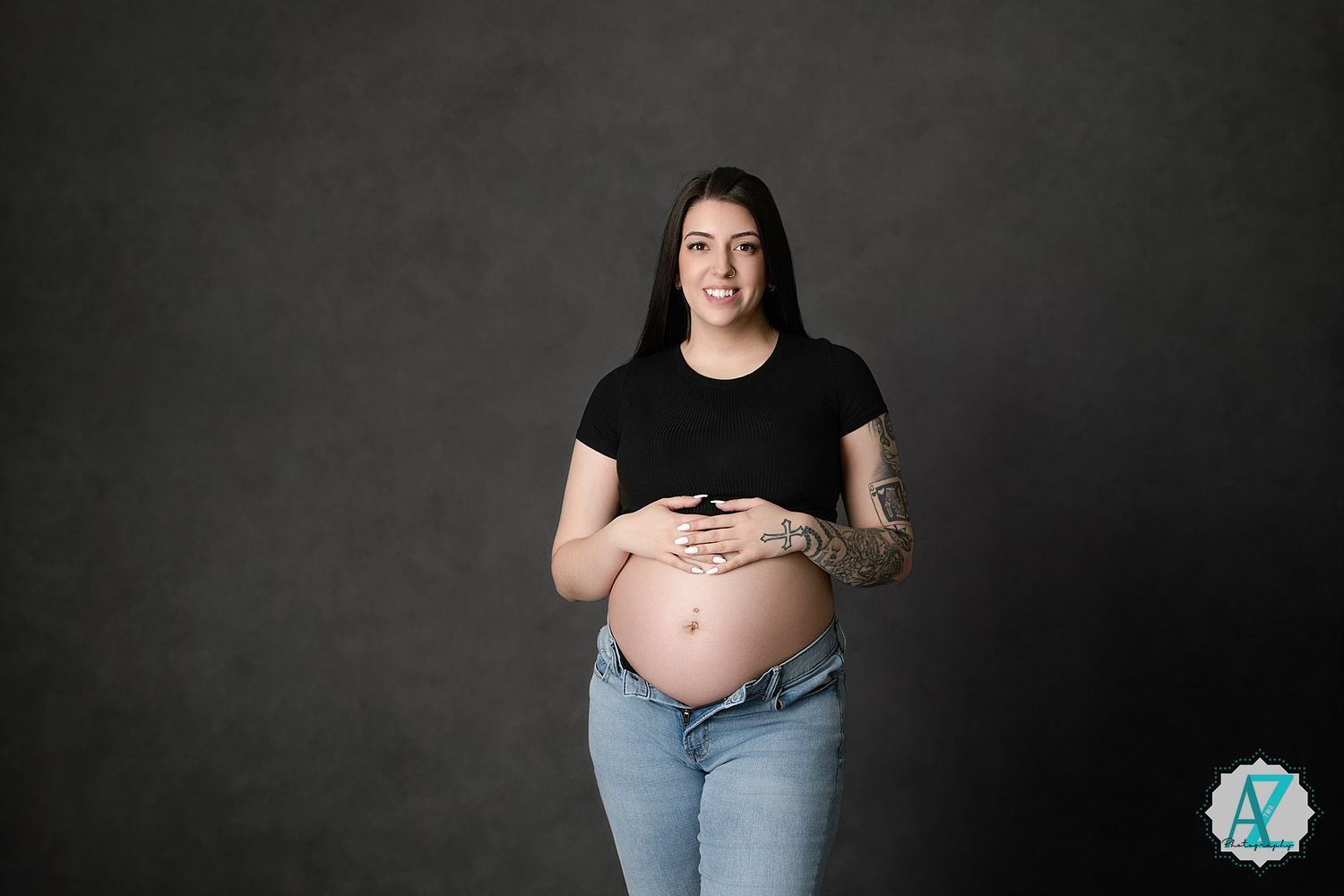 A pregnant woman in a black t-shirt and jeans poses against a dark grey backdrop during a maternity photoshoot.