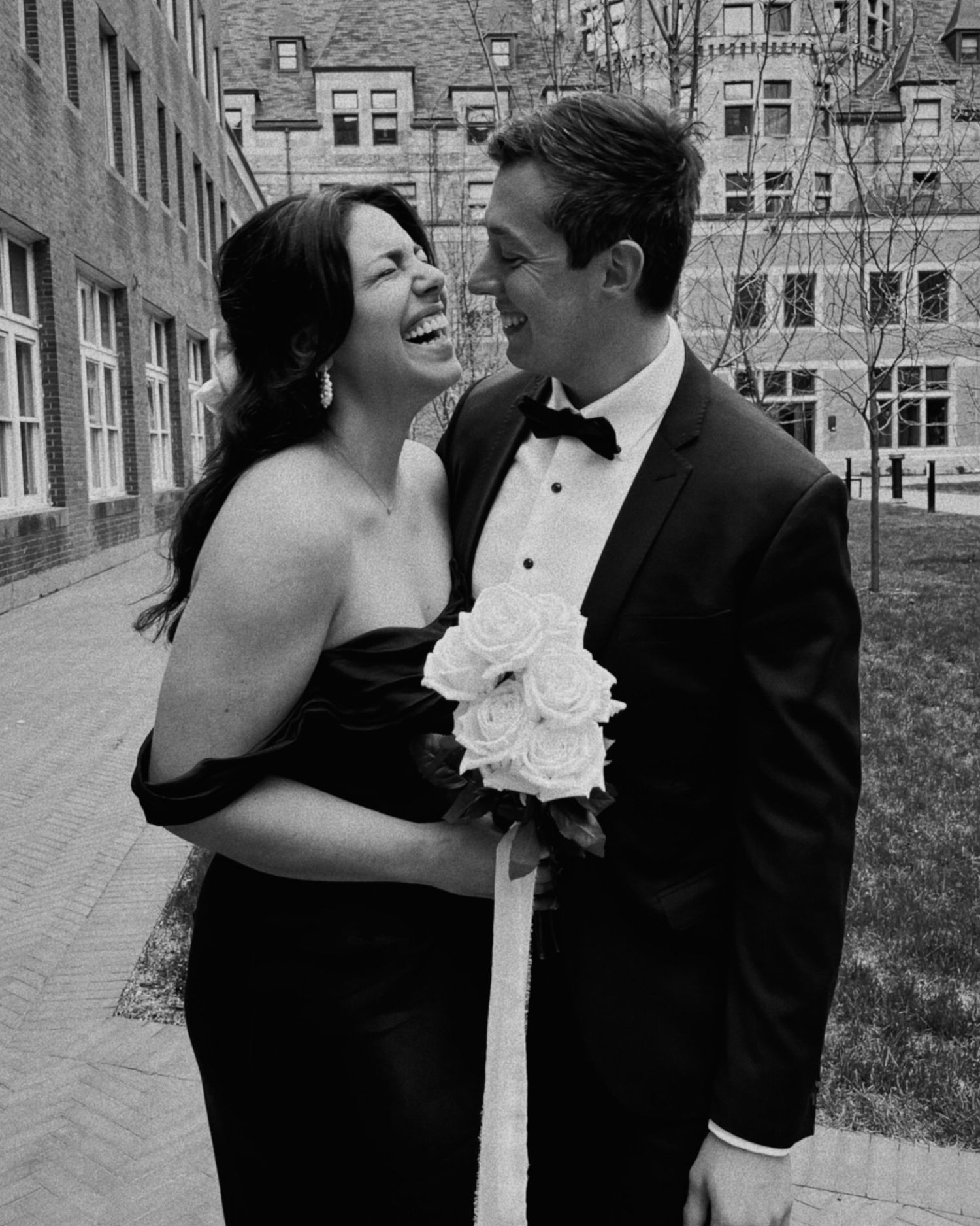 A couple shares a joyful moment in formal black attire, with white roses and brick buildings in the background.
