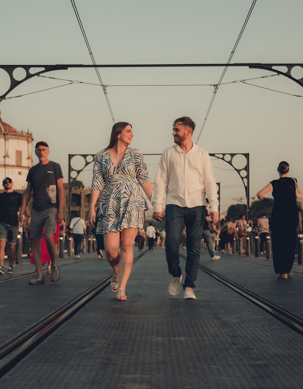 Couple walking together on a bridge walkway at sunset with other pedestrians in the background.