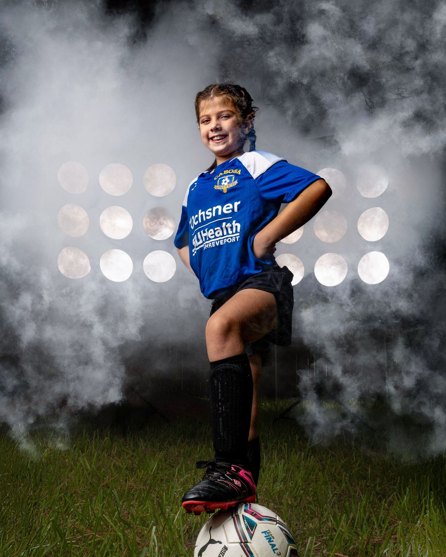 Soccer player wearing blue jersey poses dramatically with a soccer ball in dark moody lighting.