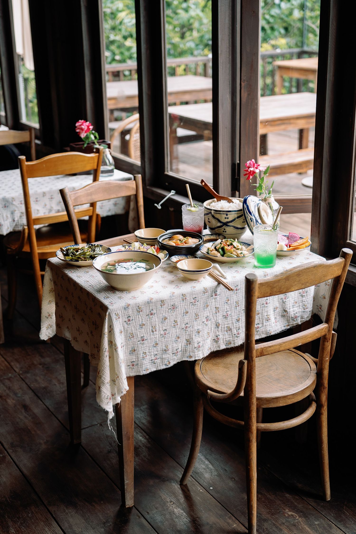 Rustic dining table set with lace tablecloth and dishes near window in cozy restaurant with pink flower centerpiece.