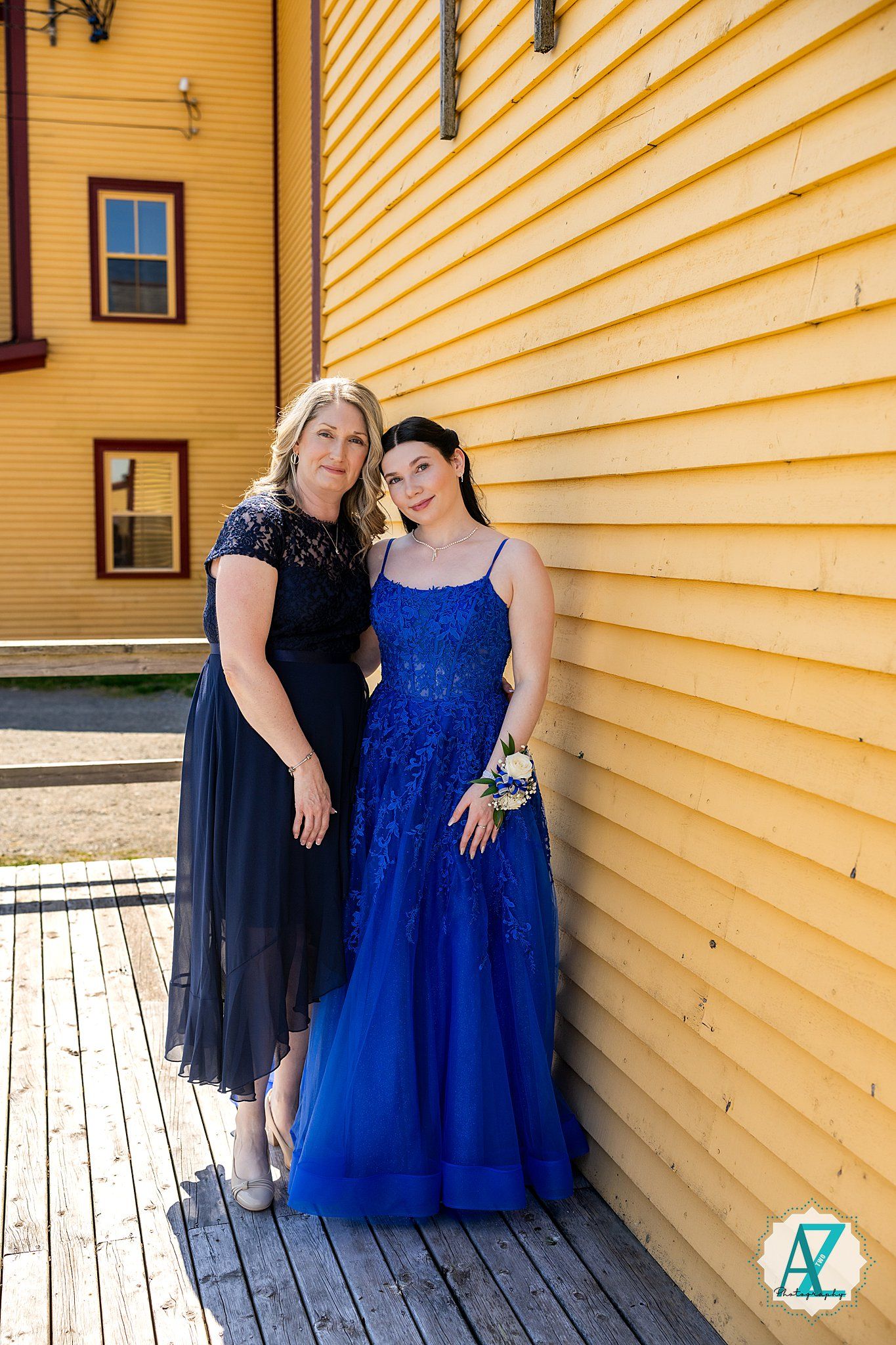 Two people in formal attire with a navy blue gown and dark dress pose against a yellow building.