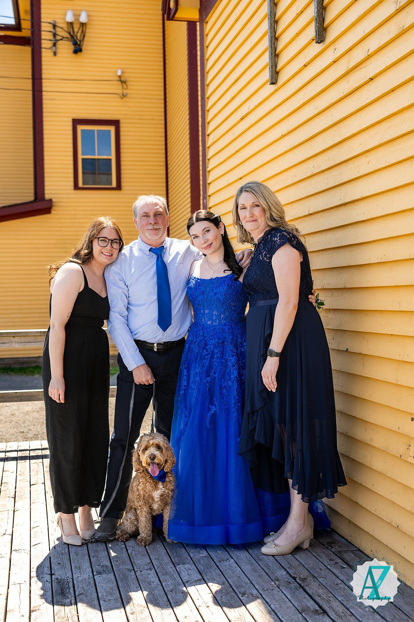 Family group in formal attire poses together with their brown dog against bright yellow house siding.