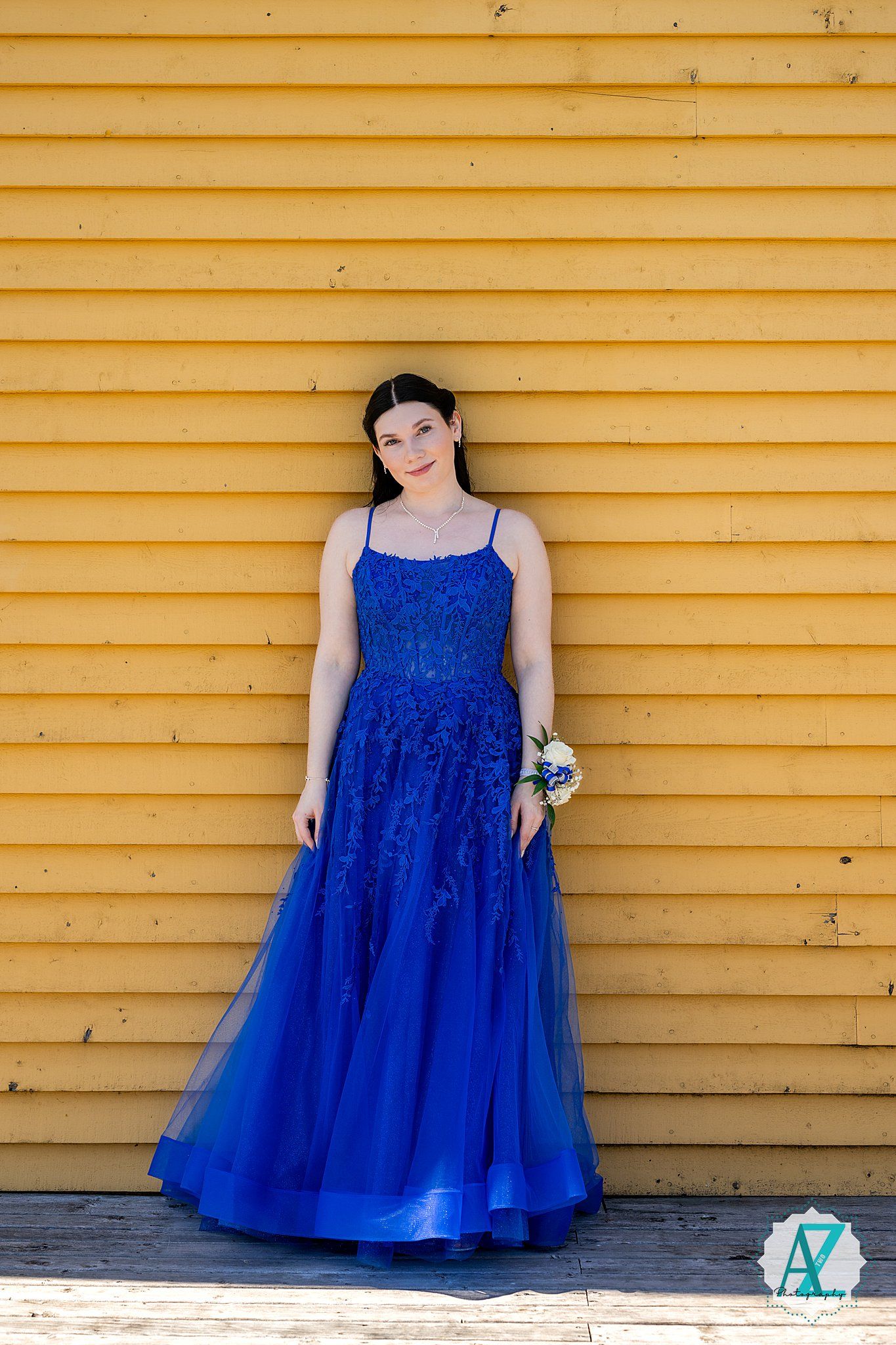 A woman in a vibrant royal blue tulle evening gown poses against a yellow wooden wall.